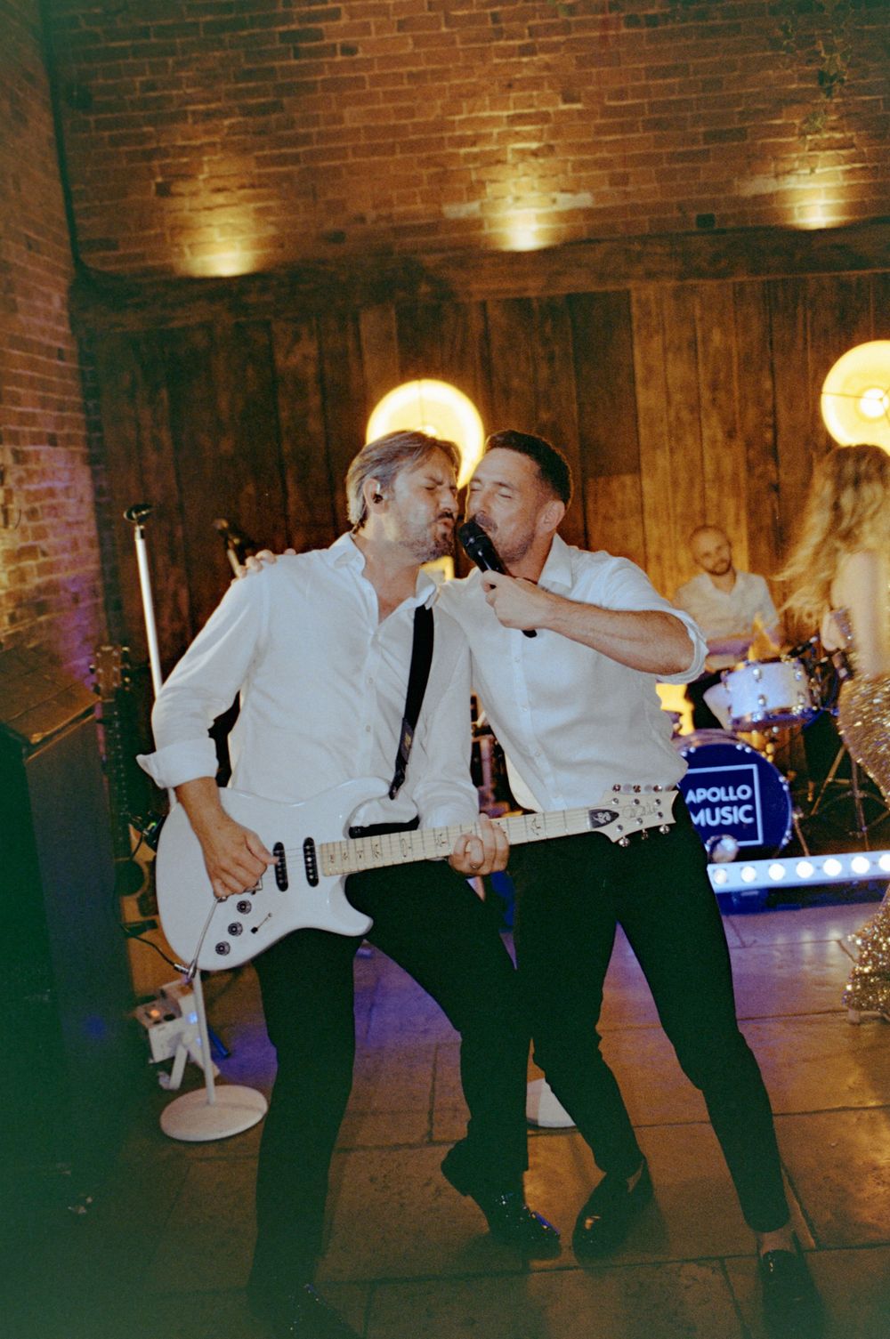 Two male musicians in white shirts perform on stage, one playing guitar, at an Apollo Music live wedding band event.