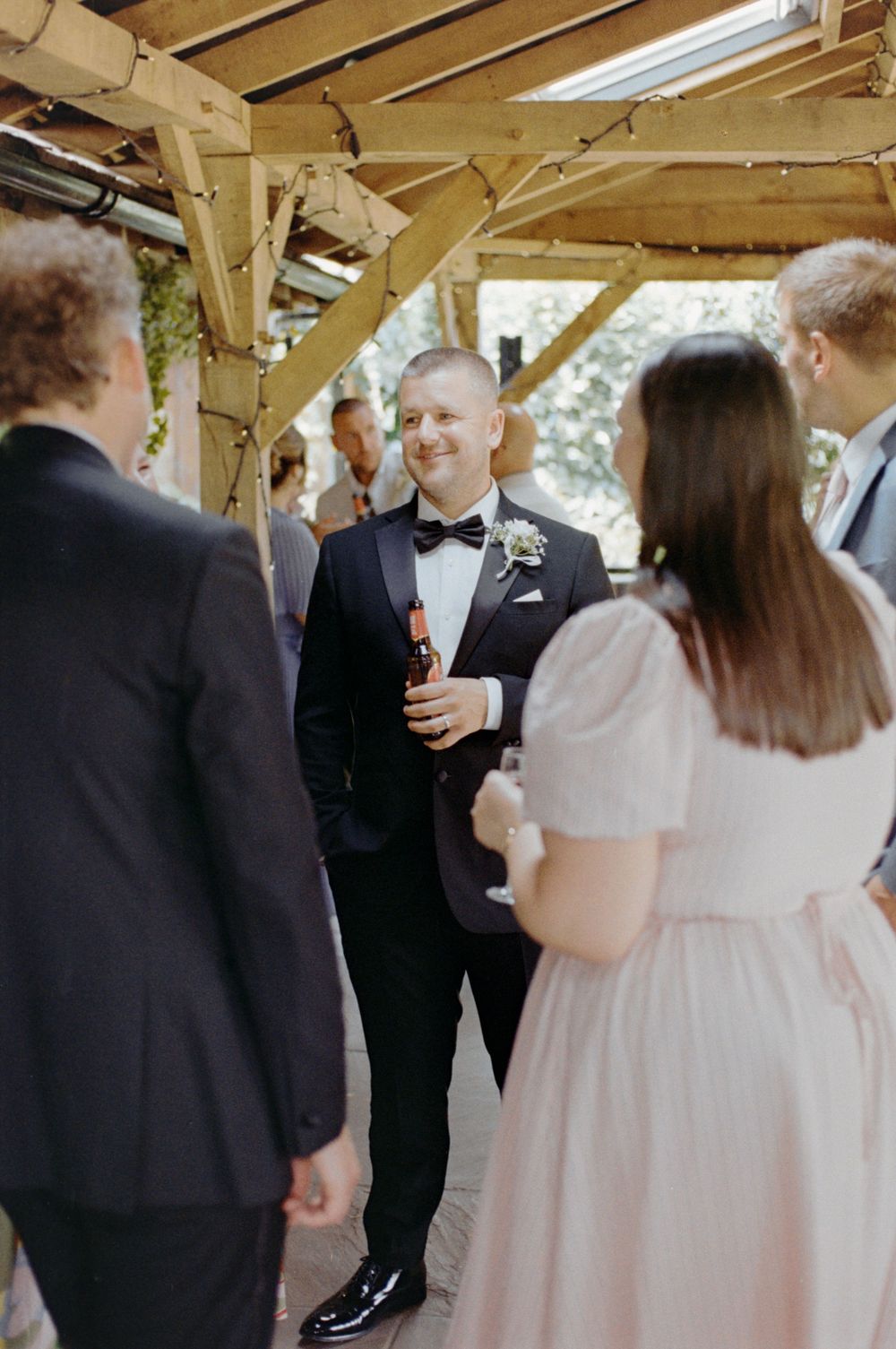 Groom in black tuxedo with bow tie holds beer bottle chatting with guests under wooden pergola at wedding reception.