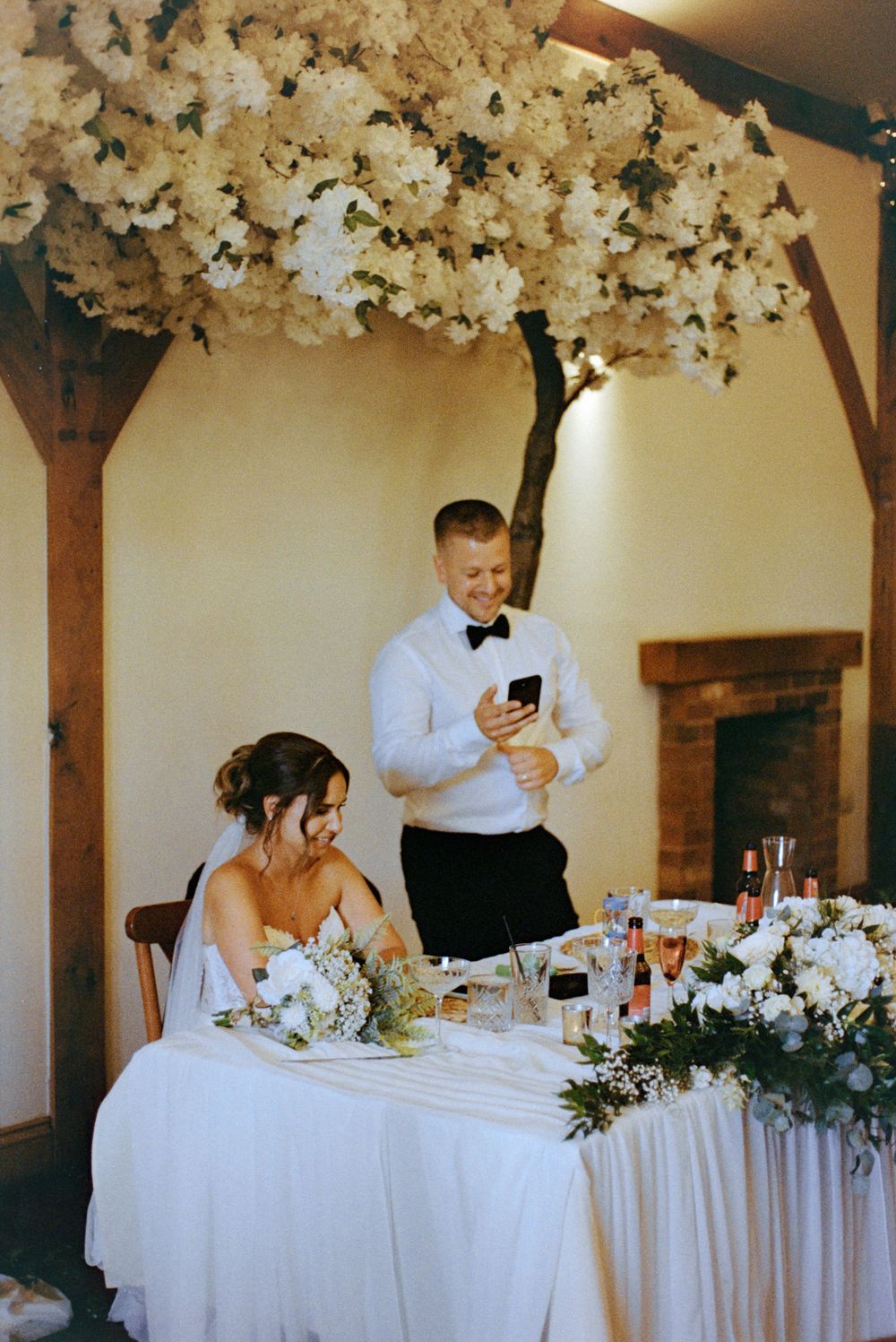 Bride seated at floral wedding reception table while groom in bow tie stands reading speech from phone.