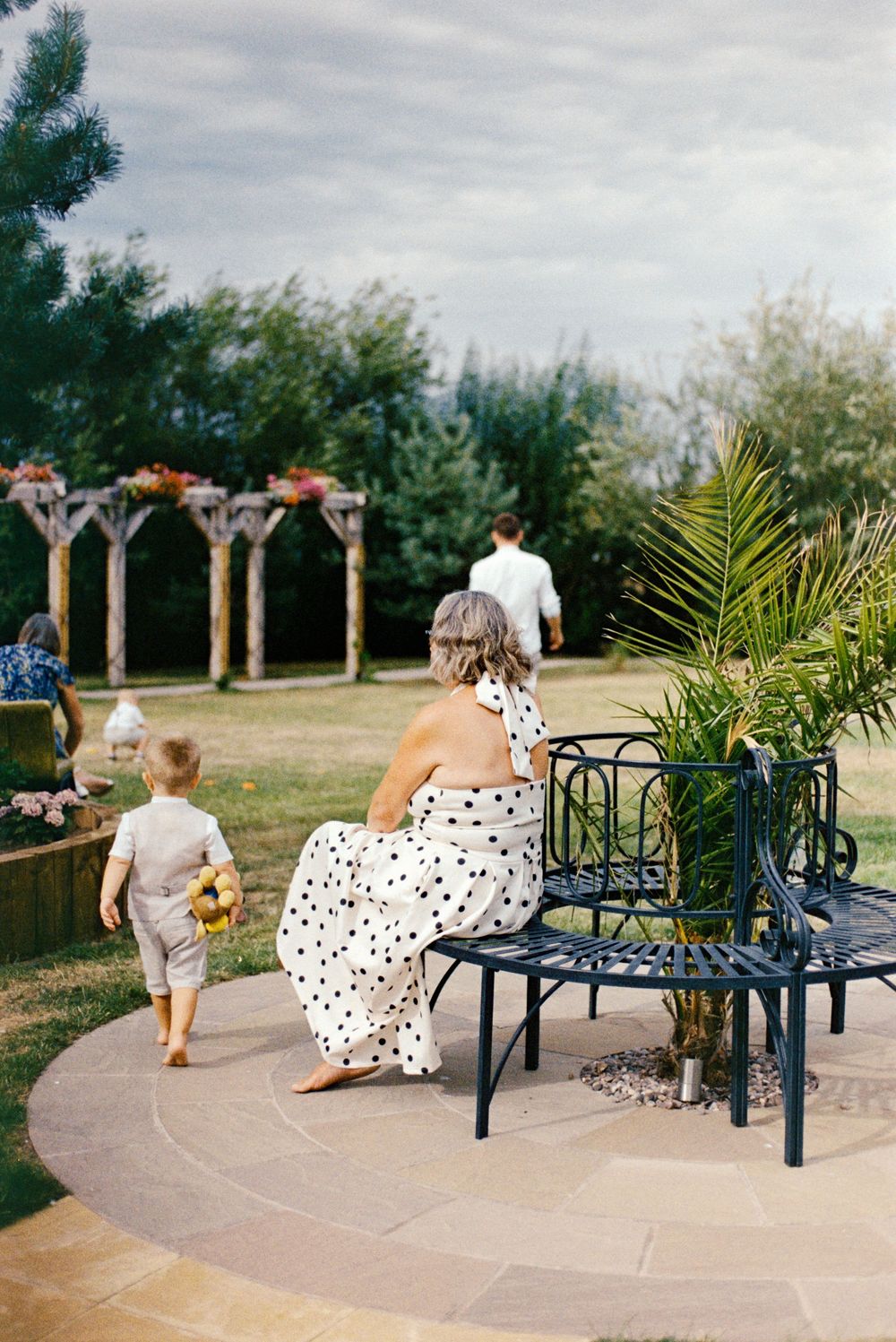 Woman in polka dot dress sits on garden bench as child in linen suit carries flowers at outdoor wedding.