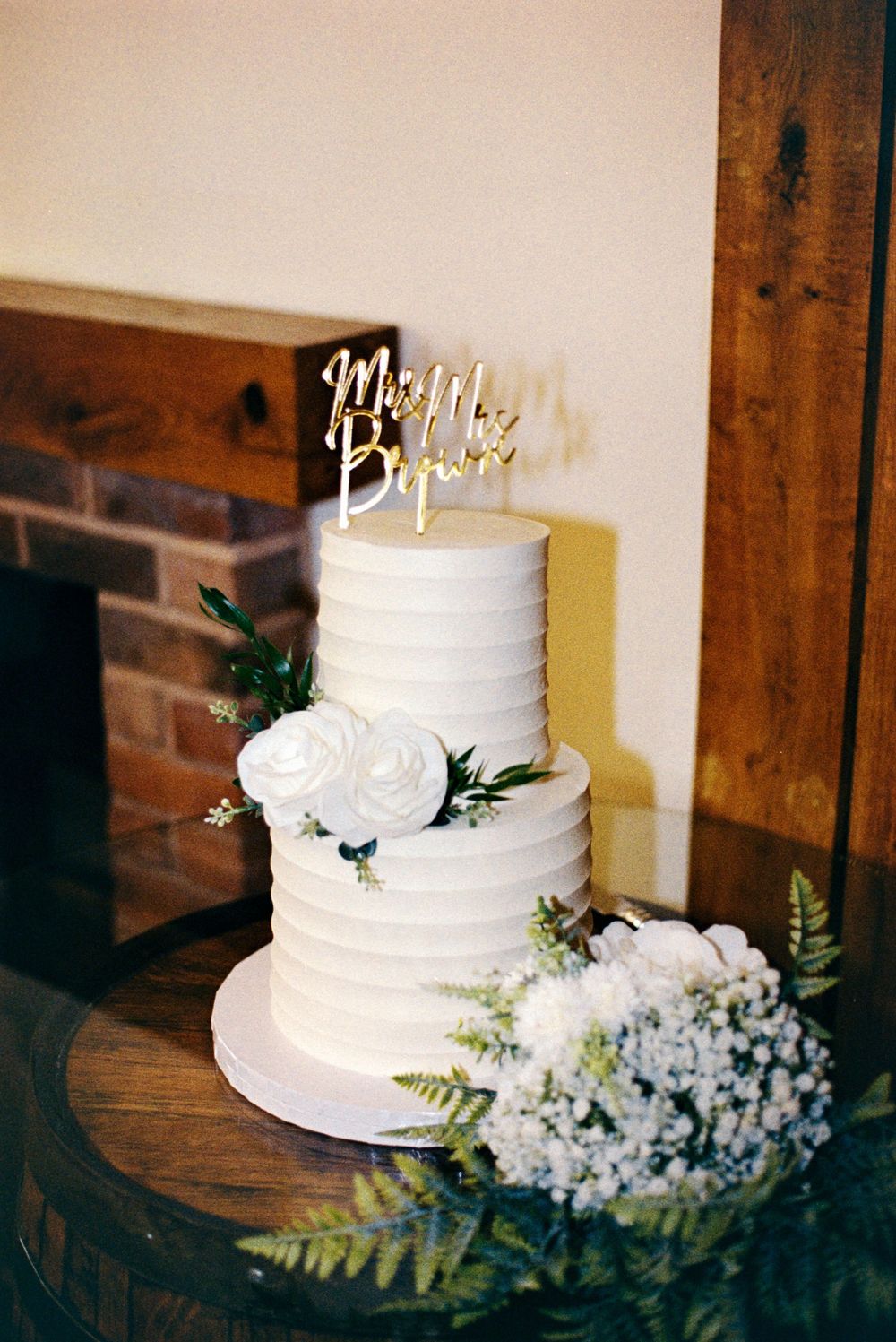 Two-tier white wedding cake with floral decor and gold Mr. & Mrs. topper, displayed near a rustic fireplace.