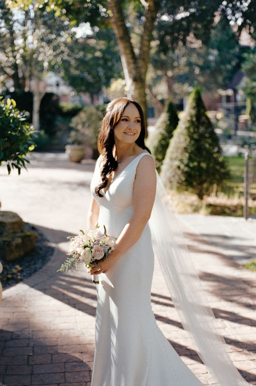 Bride in elegant white gown holding bouquet with flowing veil, posing outdoors among lush greenery on her wedding day.