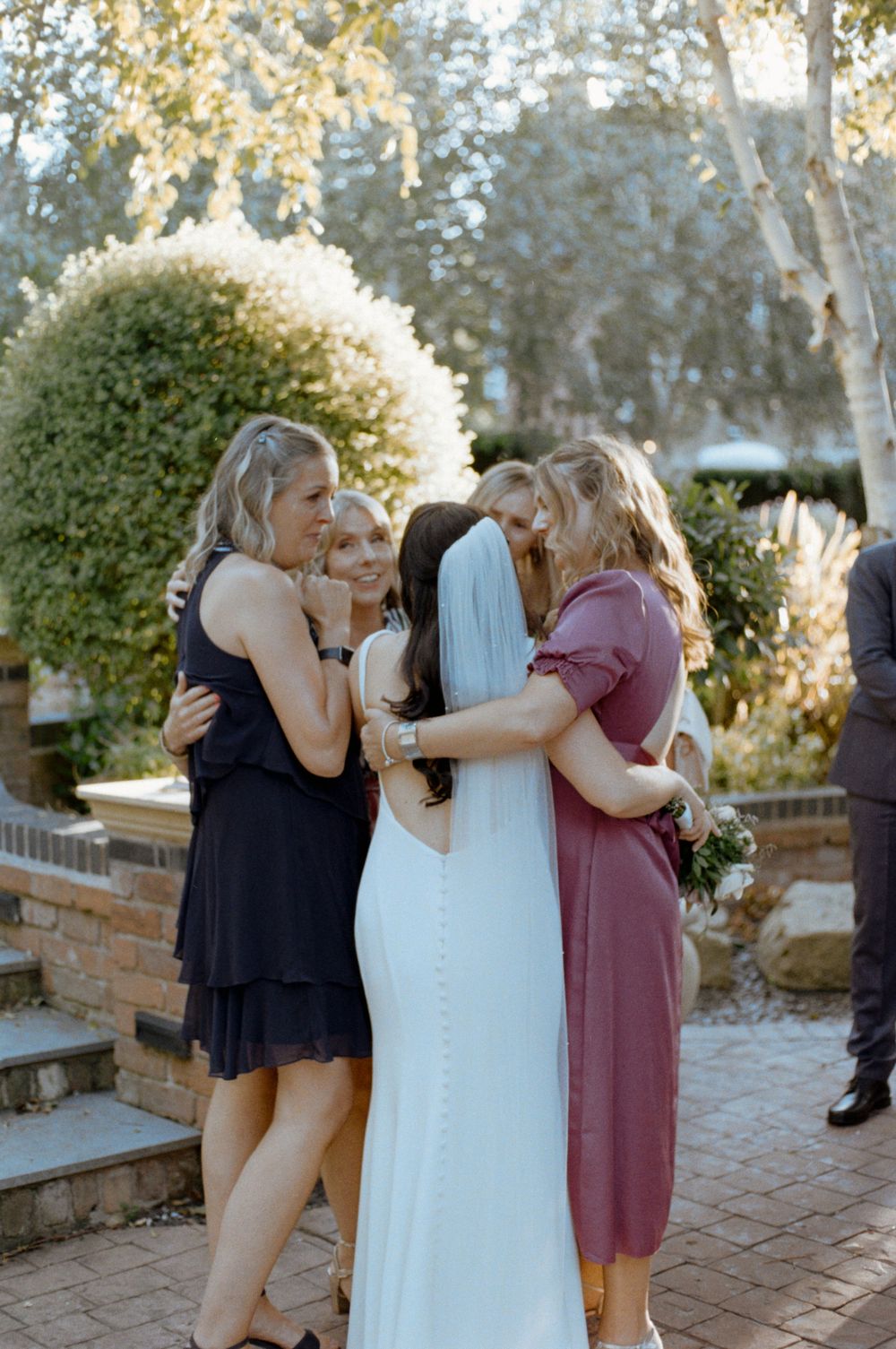 Bride in white gown and veil shares emotional group hug with three friends in garden wedding venue.