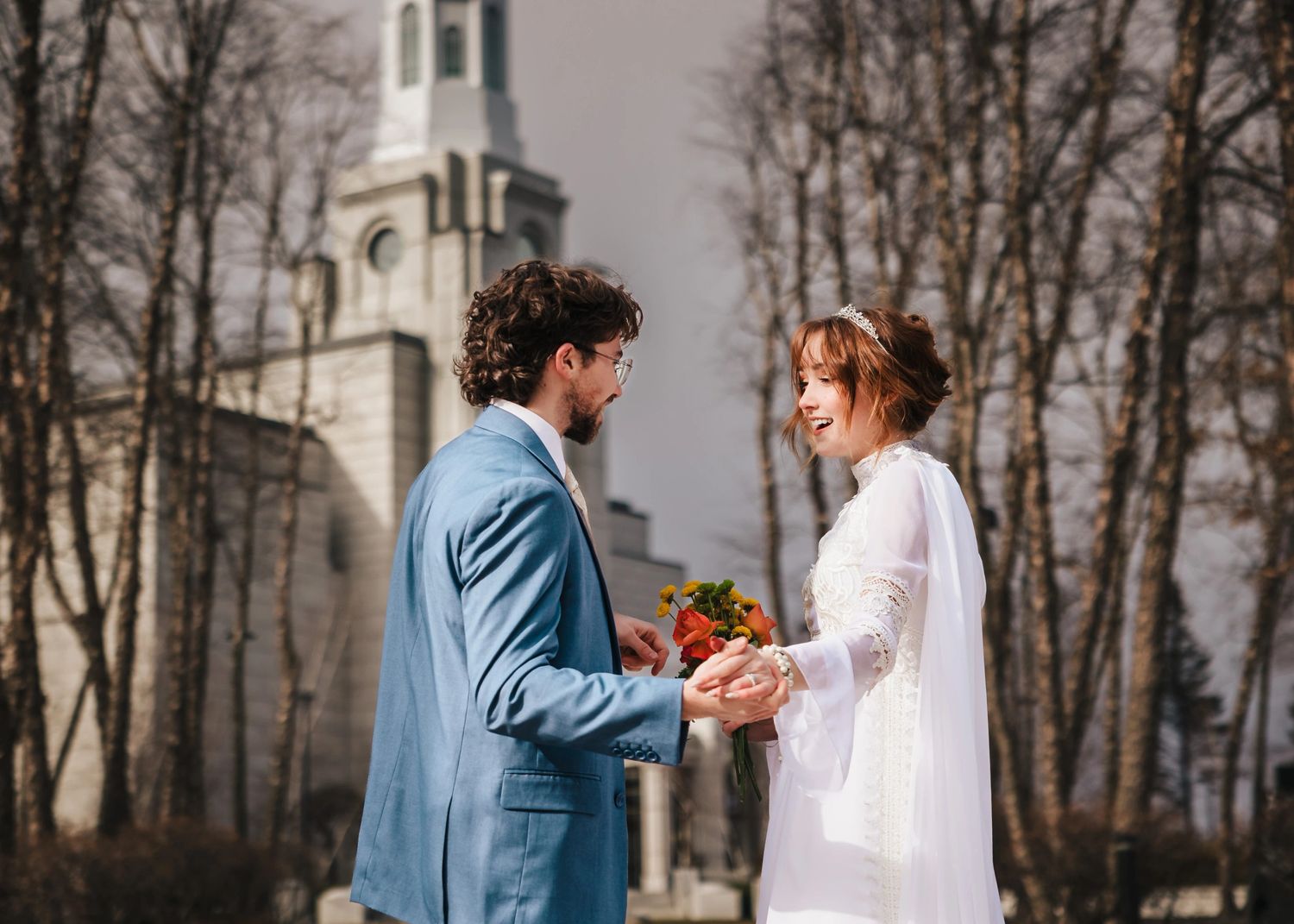 Couple exchanging vows or rings during an outdoor ceremony, facing each other with a church visible behind them.