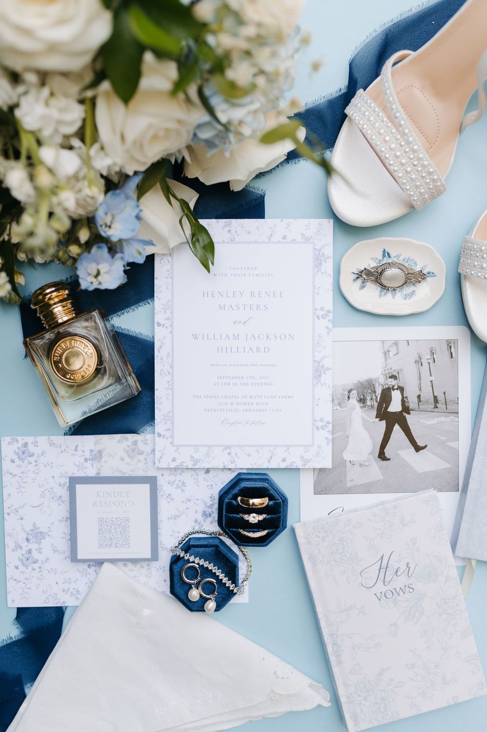 Elegant wedding flat lay with blue velvet ring box, gold rings, floral invitation suite, perfume bottle, bridal heels, and white flowers.