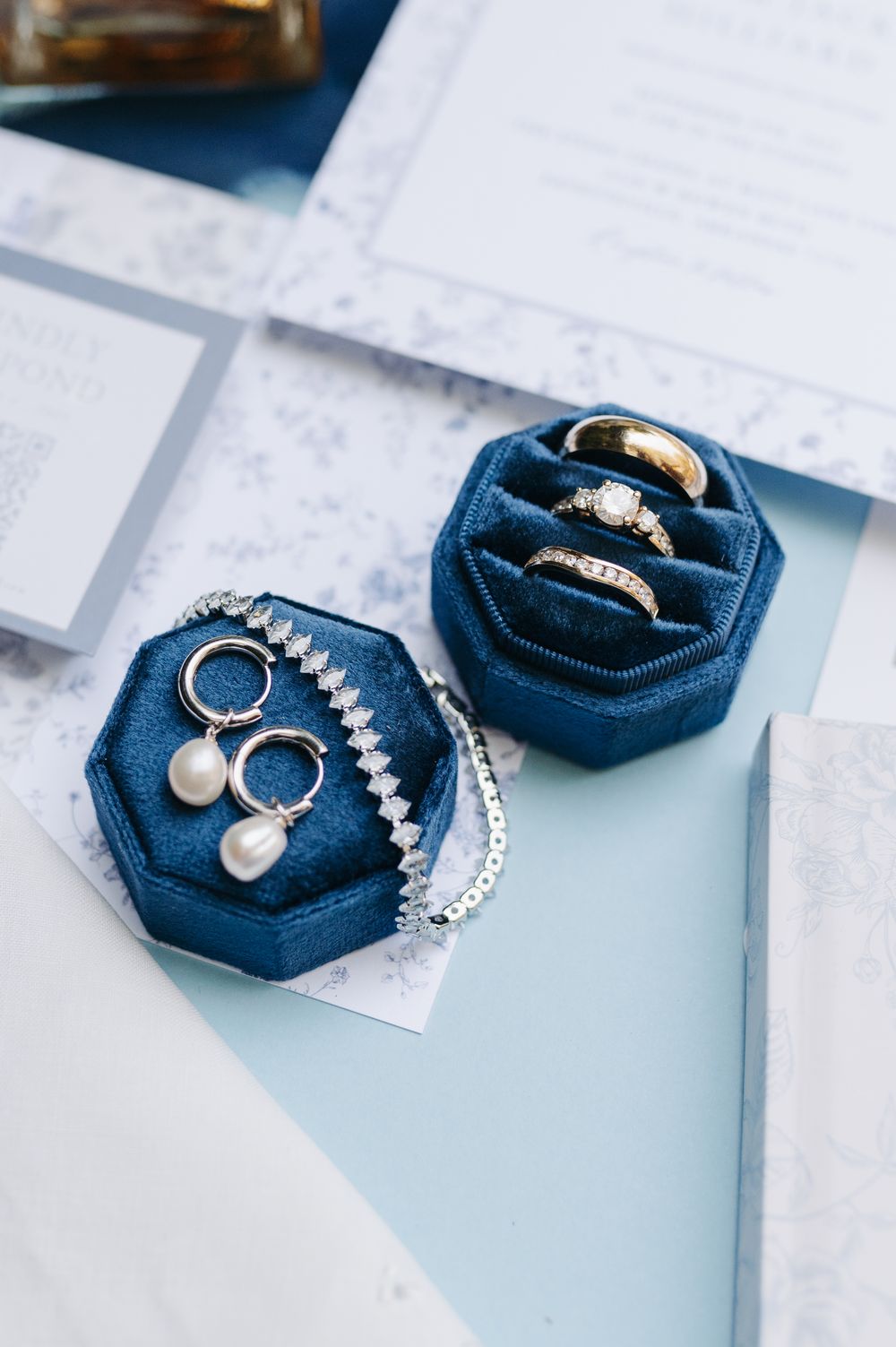 Blue velvet hexagonal jewelry boxes displaying pearl earrings, diamond bracelet, and gold wedding rings on elegant stationery.