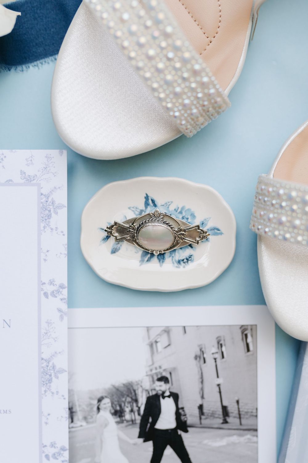 White pearl wedding sandals beside a floral ring dish and black-and-white couple photo on a blue background.