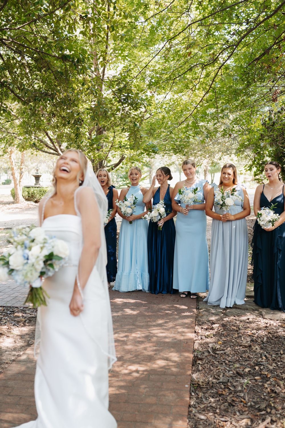 Bride laughing joyfully with bridesmaids in blue dresses holding white bouquets outdoors under green trees.