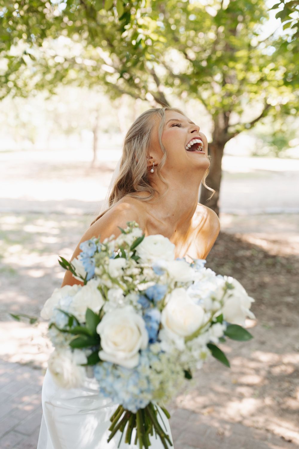 Laughing bride in strapless white gown holding blue and white floral bouquet outdoors under green trees.
