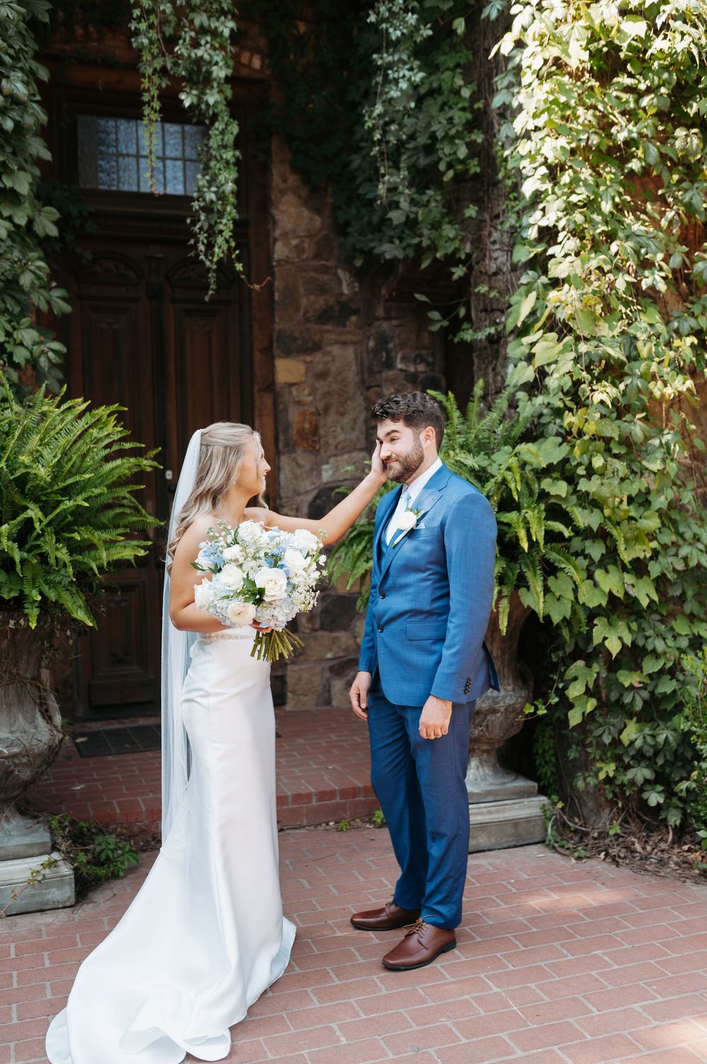 Bride in white gown touches groom's face during first look outside ivy-covered stone building.