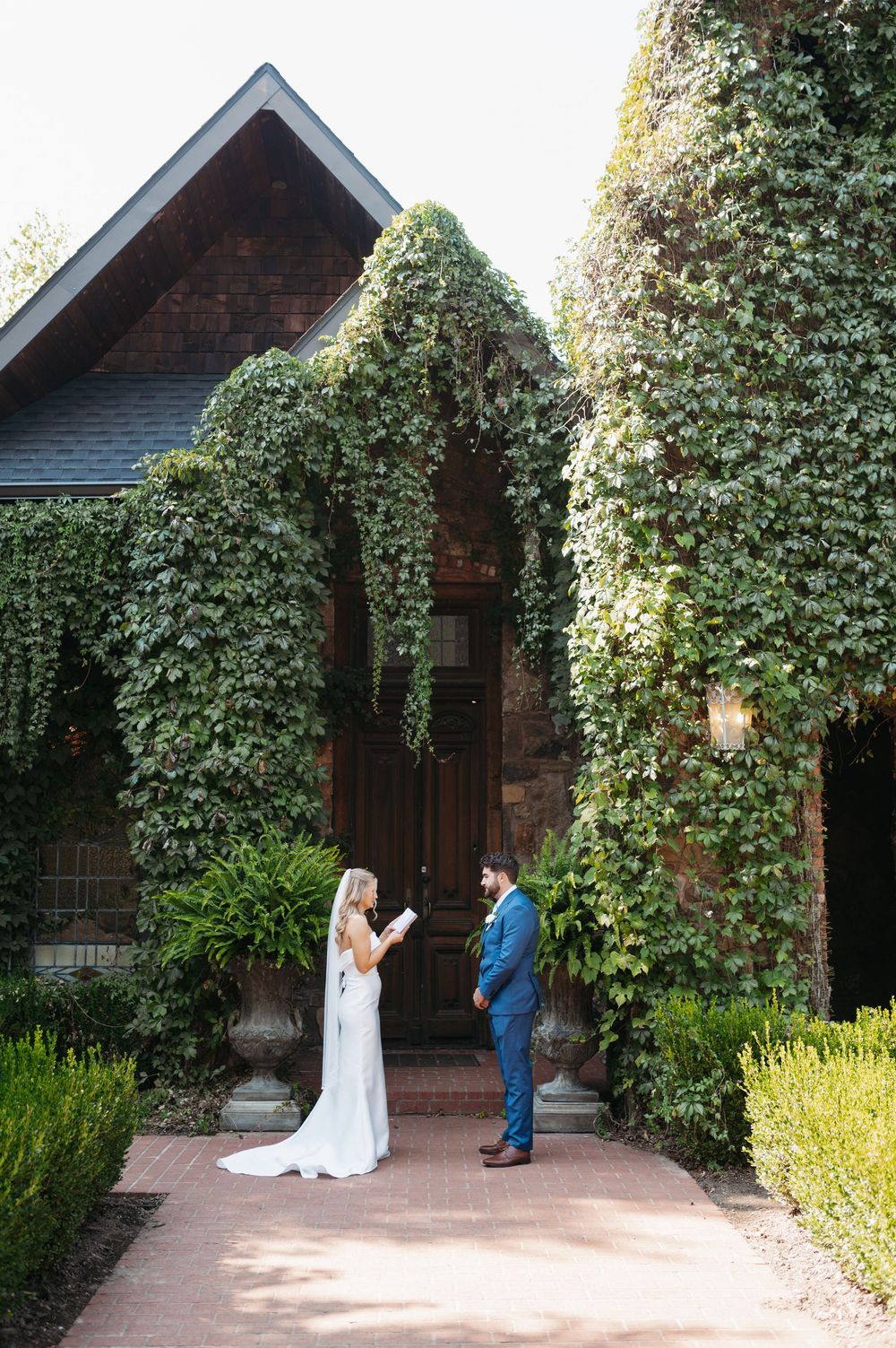 Bride reads vows to groom outside ivy-covered stone chapel during outdoor wedding ceremony.