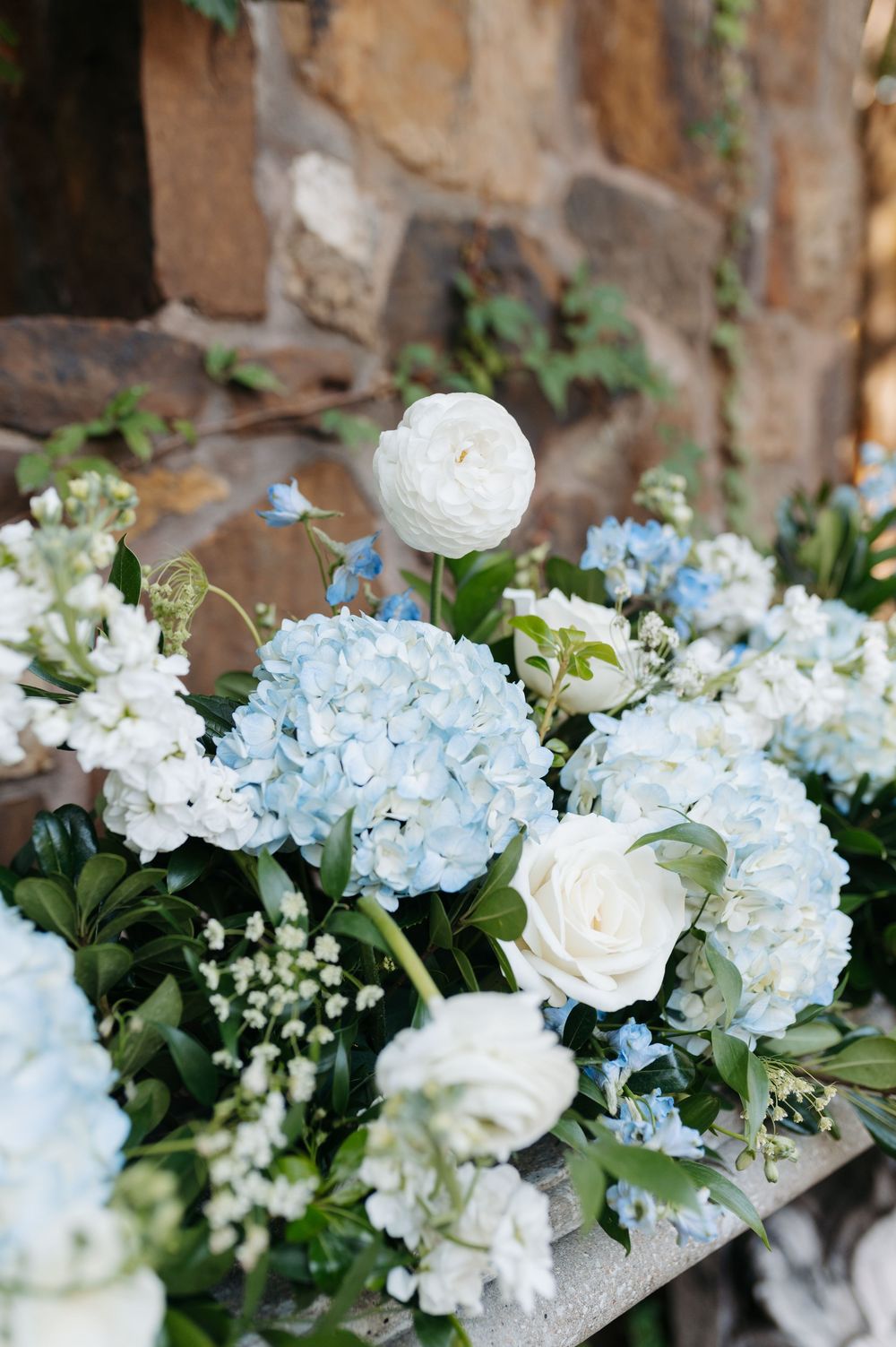 Wedding floral arrangement of blue hydrangeas, white ranunculus, and roses against stone wall.