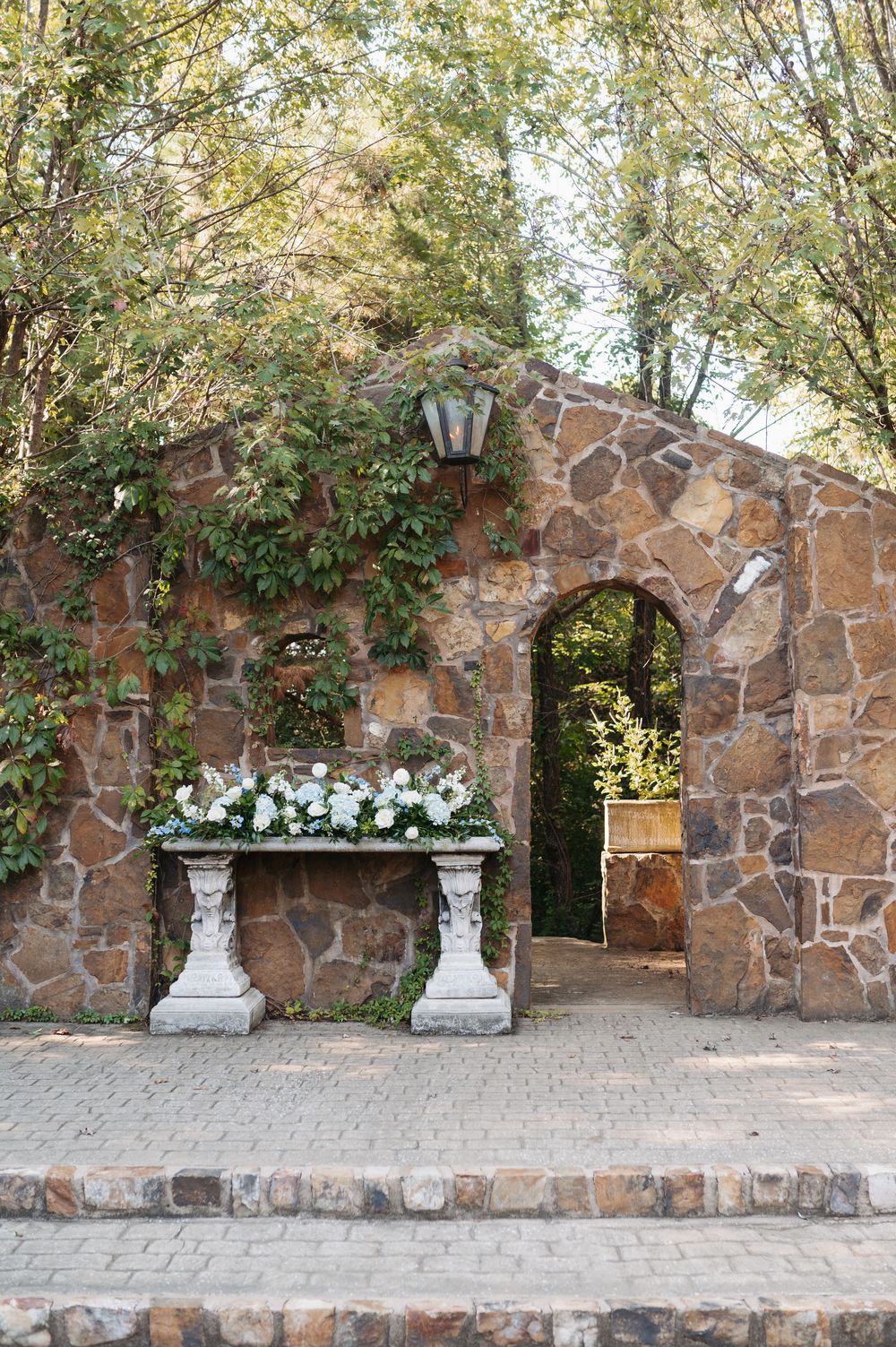 Stone arch garden wall with white floral arrangement on ornate pedestals at wedding venue.