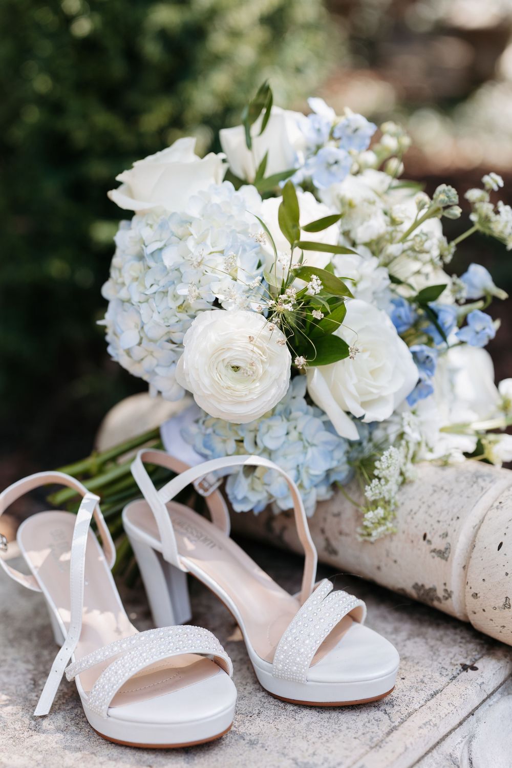 White bridal heels beside a bouquet of blue hydrangeas and white roses on stone steps.