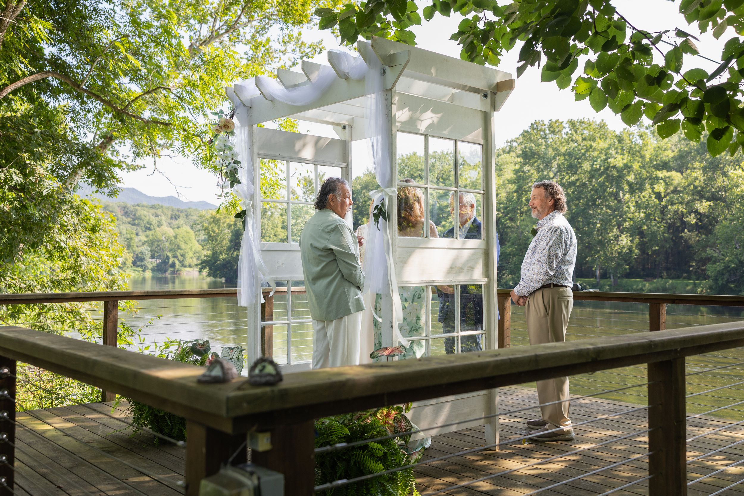 Outdoor wedding ceremony on a wooden deck by a lake, with couple under white pergola surrounded by lush greenery.