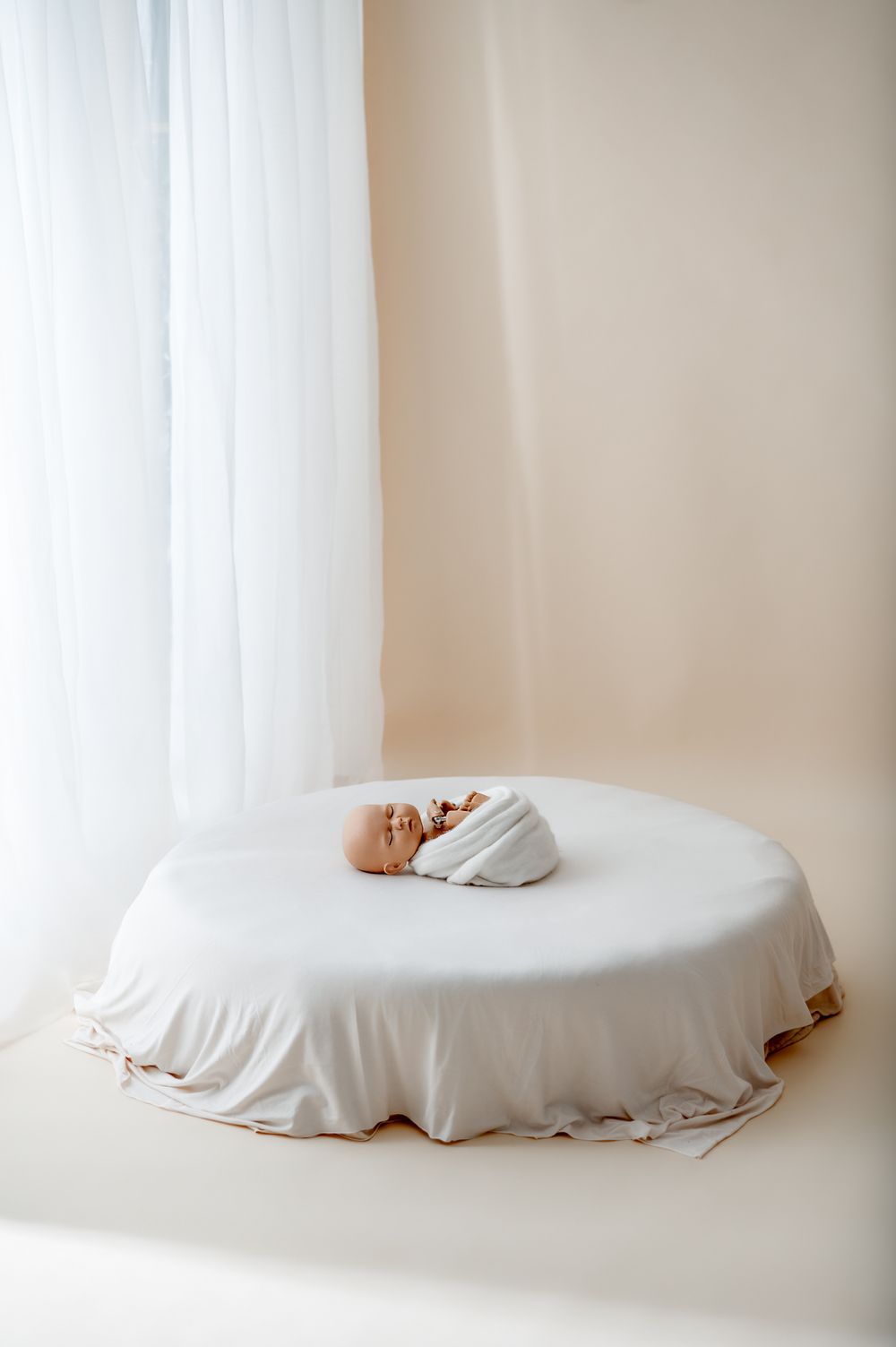 Newborn baby swaddled in white blanket sleeping peacefully on a round white bed near sheer curtains.