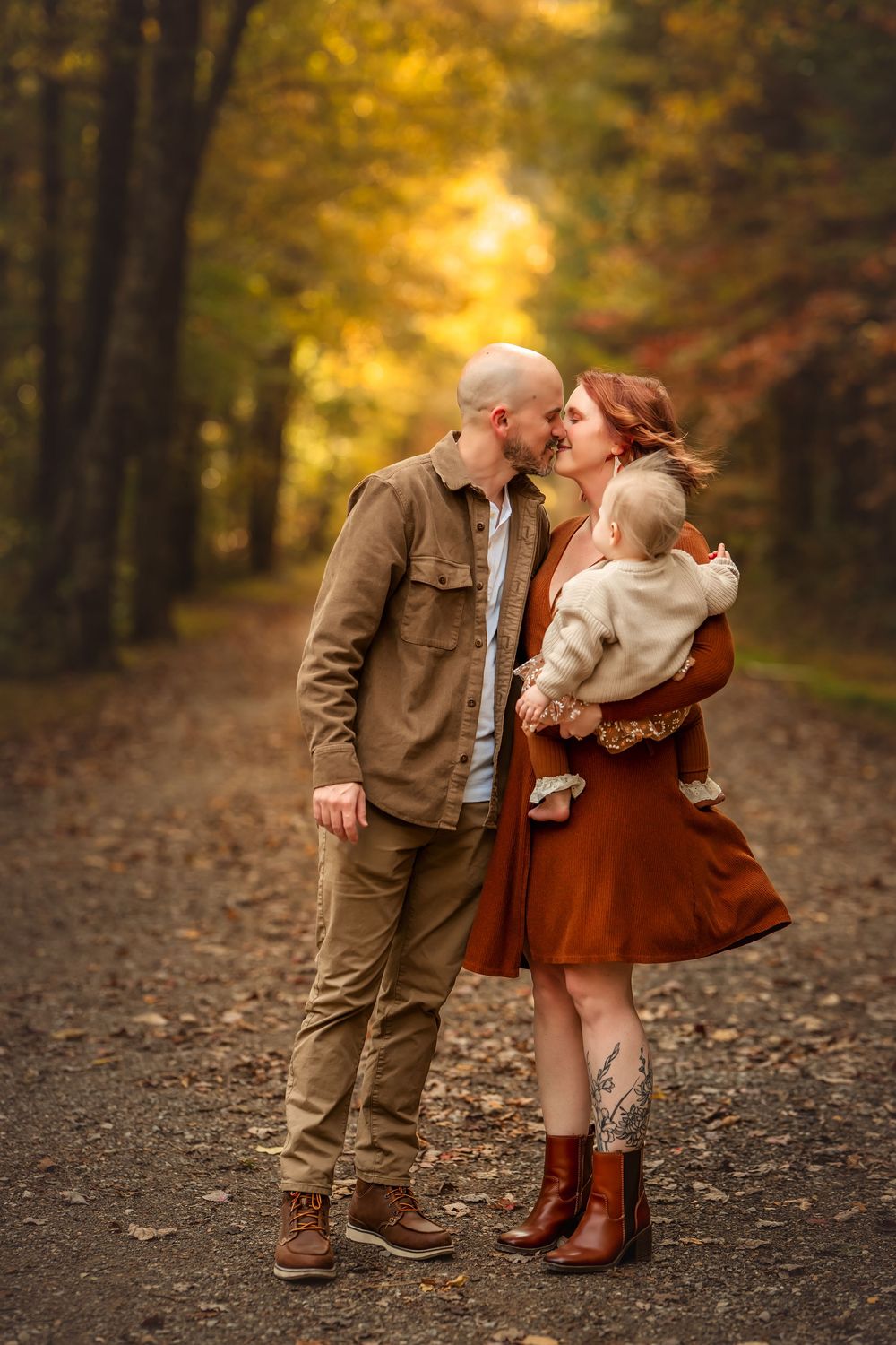 A couple shares a kiss on an autumn forest path while holding their baby, dressed in warm earth tones.