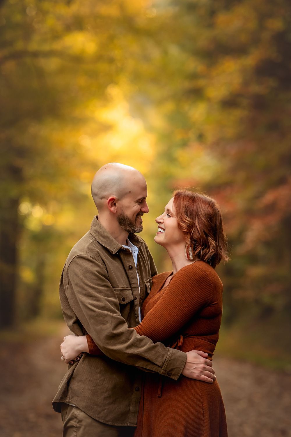 Couple embracing and smiling at each other on a forest path surrounded by vibrant autumn foliage.