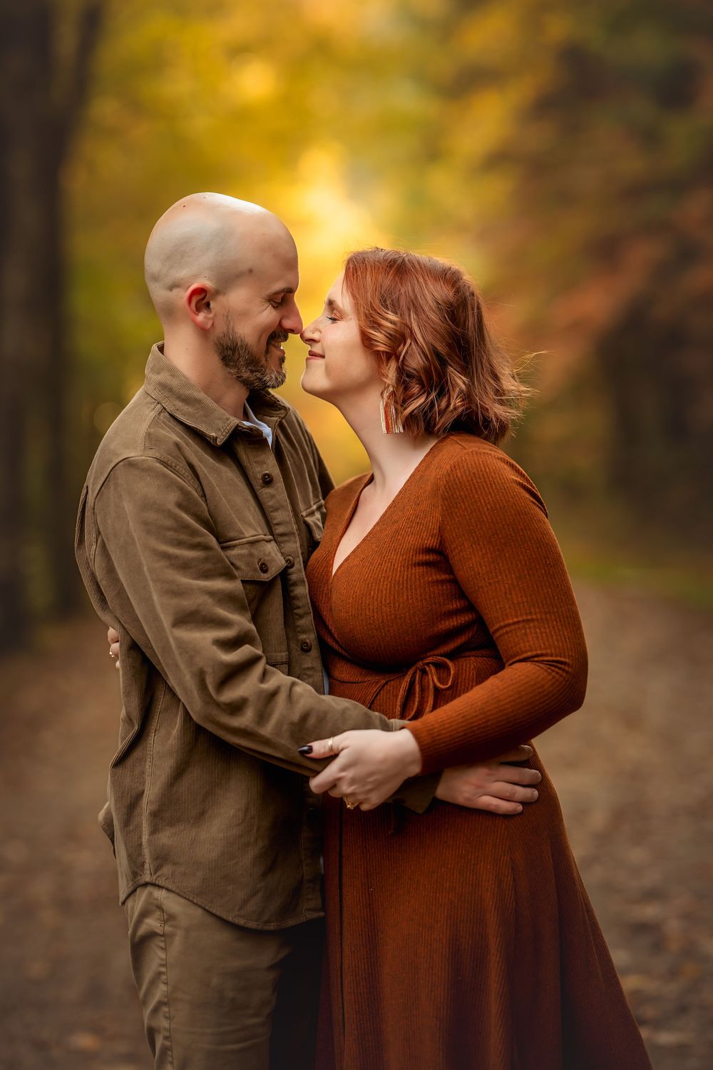 A couple shares a romantic moment on a forest path surrounded by vibrant autumn foliage in warm golden light.
