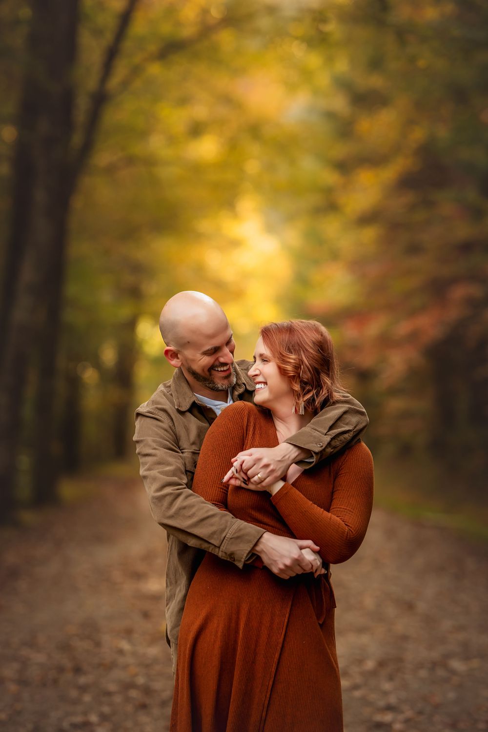 Couple embracing and smiling on a tree-lined path surrounded by vibrant autumn foliage.