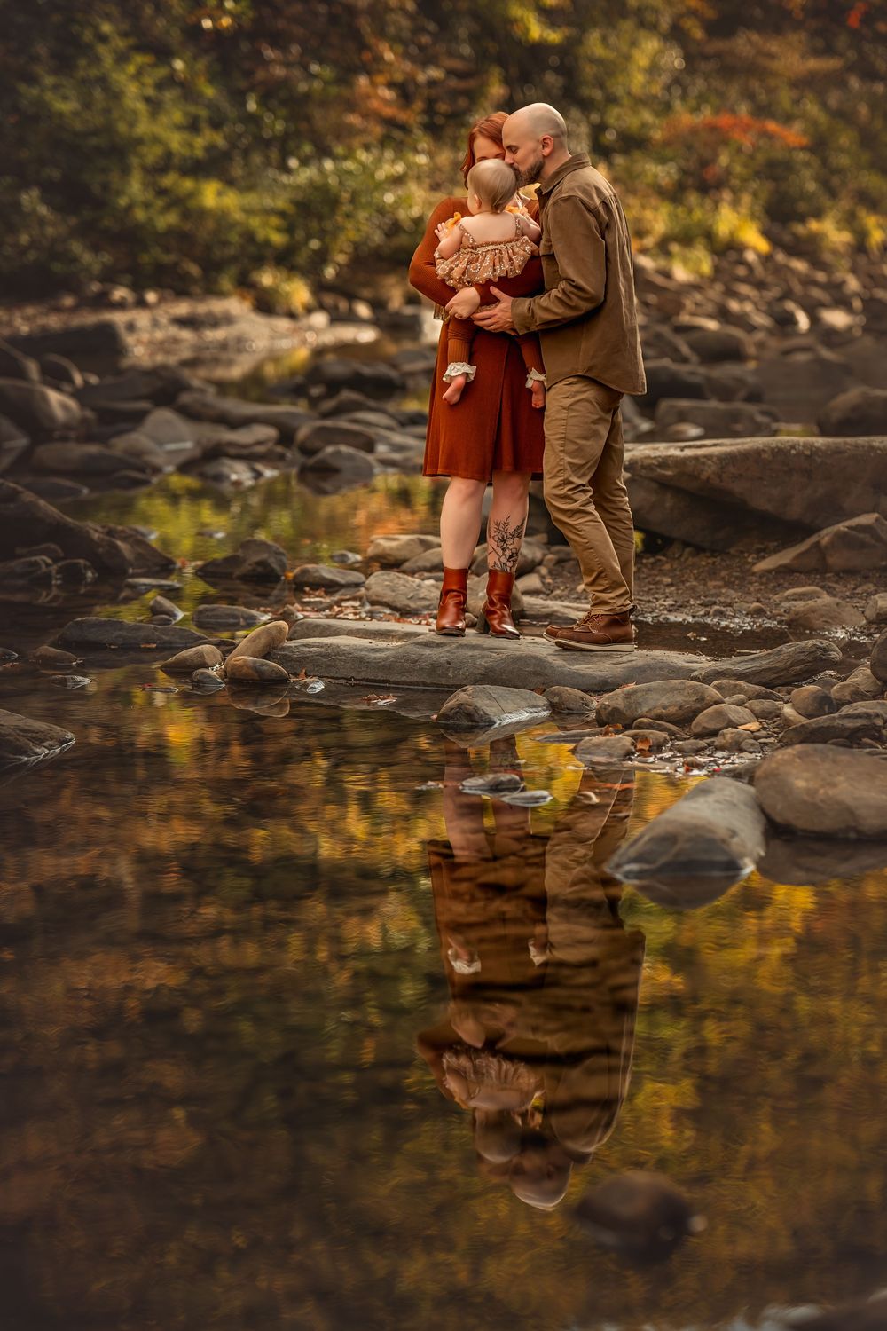 Couple holding a baby by a rocky stream with fall foliage reflection in water.