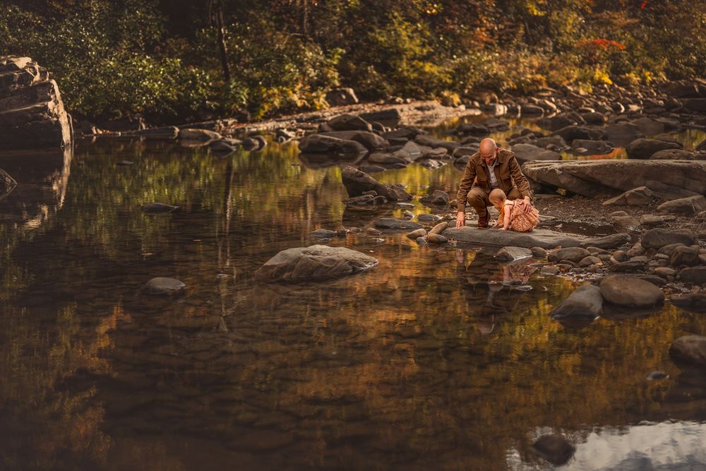 Two people sit on rocks by a calm reflective river surrounded by autumn foliage in a serene forest setting.