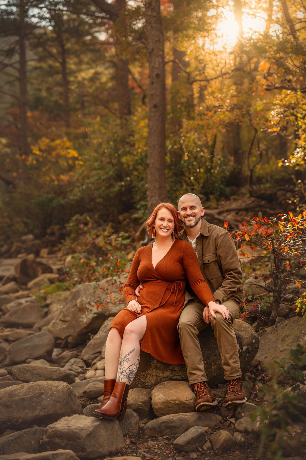 Couple sitting on rocks in autumn forest, woman in rust dress and boots, man in olive jacket, golden sunlight filtering through trees.