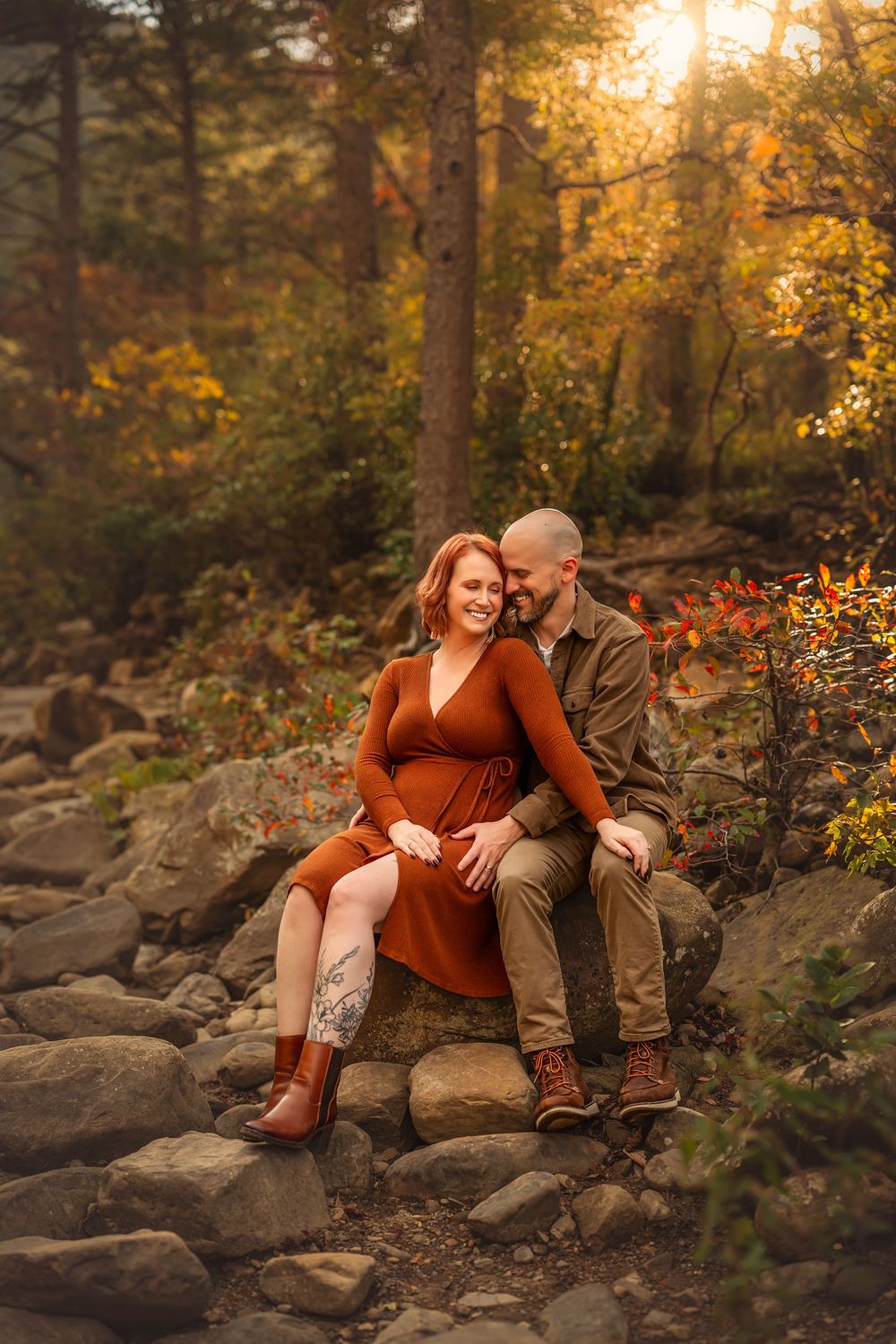 Couple sits on rocks in autumn forest, woman in rust dress, man in olive jacket, golden sunlight filtering through trees.