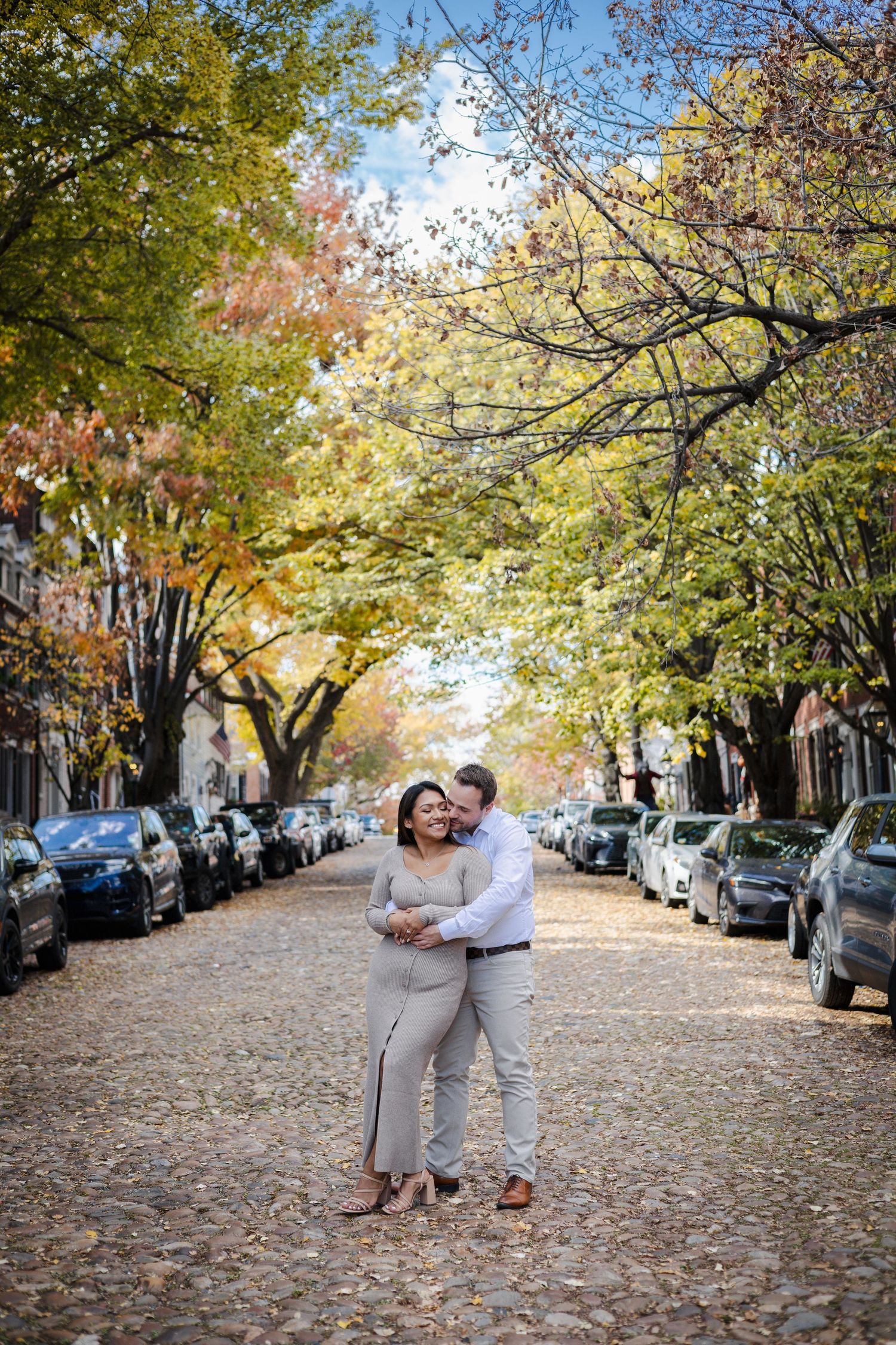 Couple embraces on a cobblestone street lined with autumn trees and parked cars in a charming urban neighborhood.