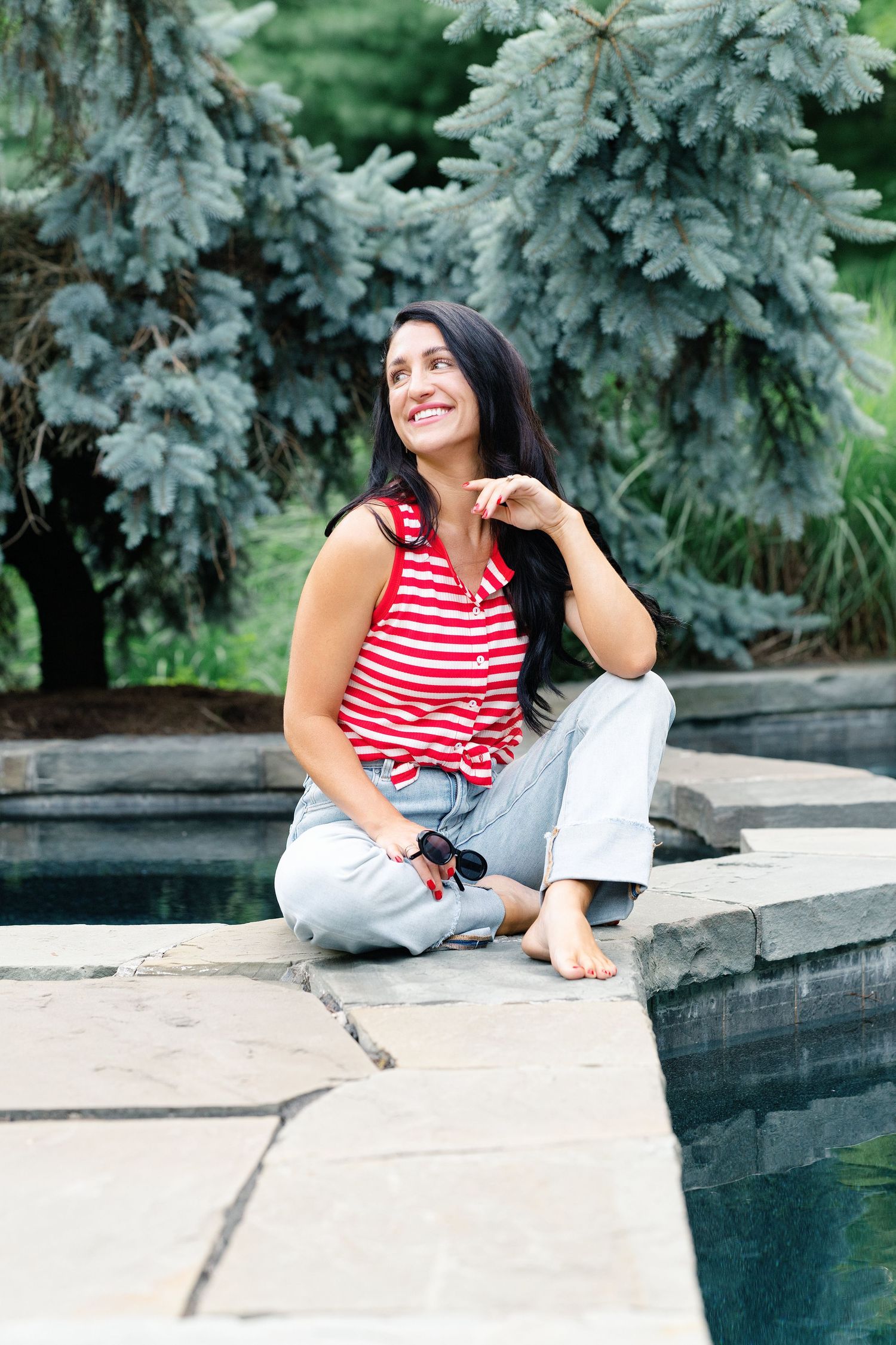Laughing blonde woman sitting cross-legged on stone patio in blue striped shirt and jeans near evergreen trees.
