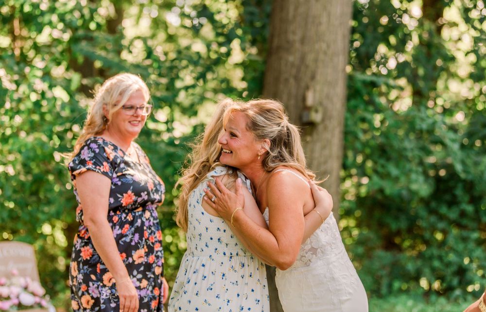 Two women hugging warmly outdoors at a garden wedding while a smiling older woman watches nearby.