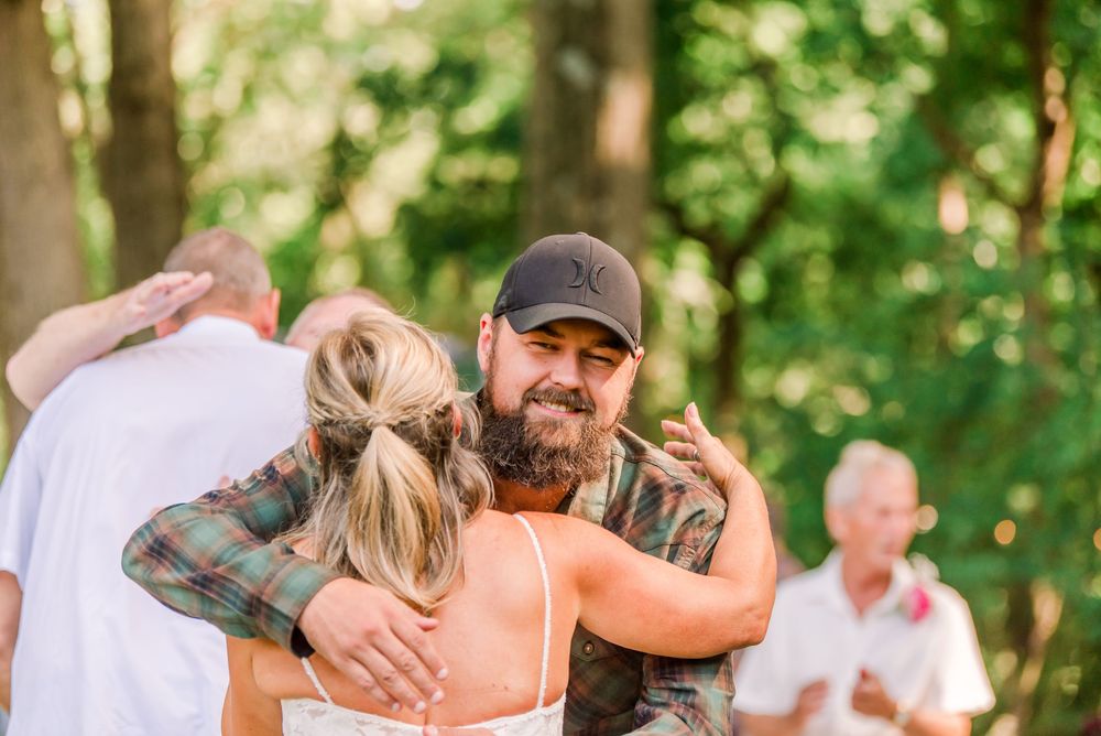 Man in plaid shirt and cap hugging a woman outdoors at a woodland wedding celebration.