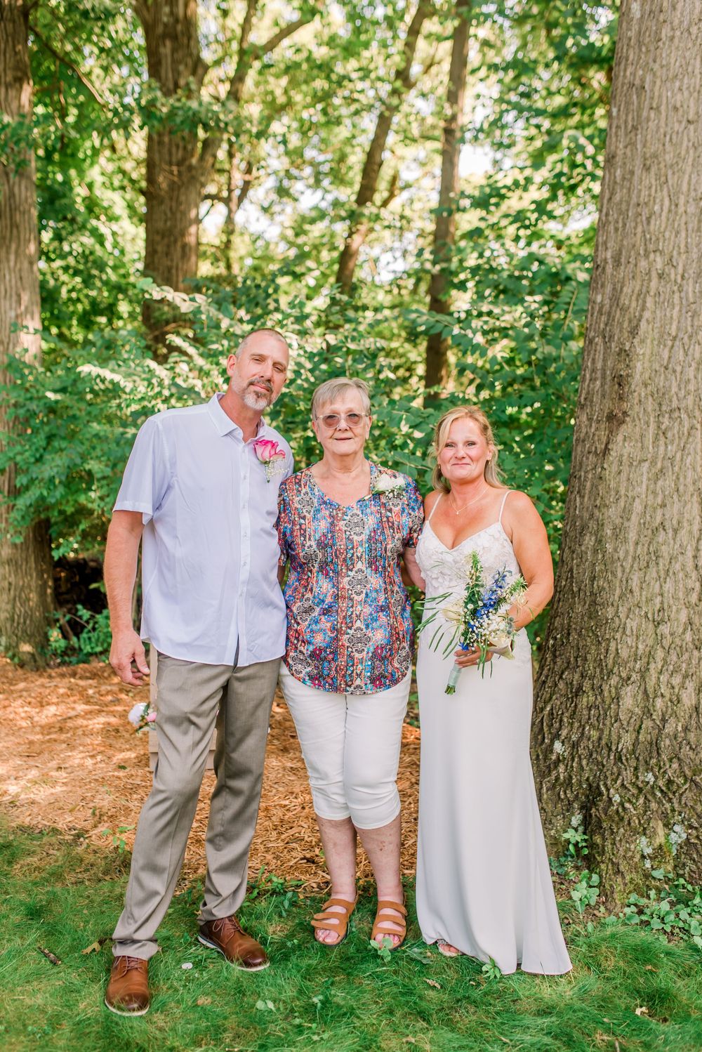 A bride in white dress holds bouquet beside groom and older woman guest outdoors near large trees.