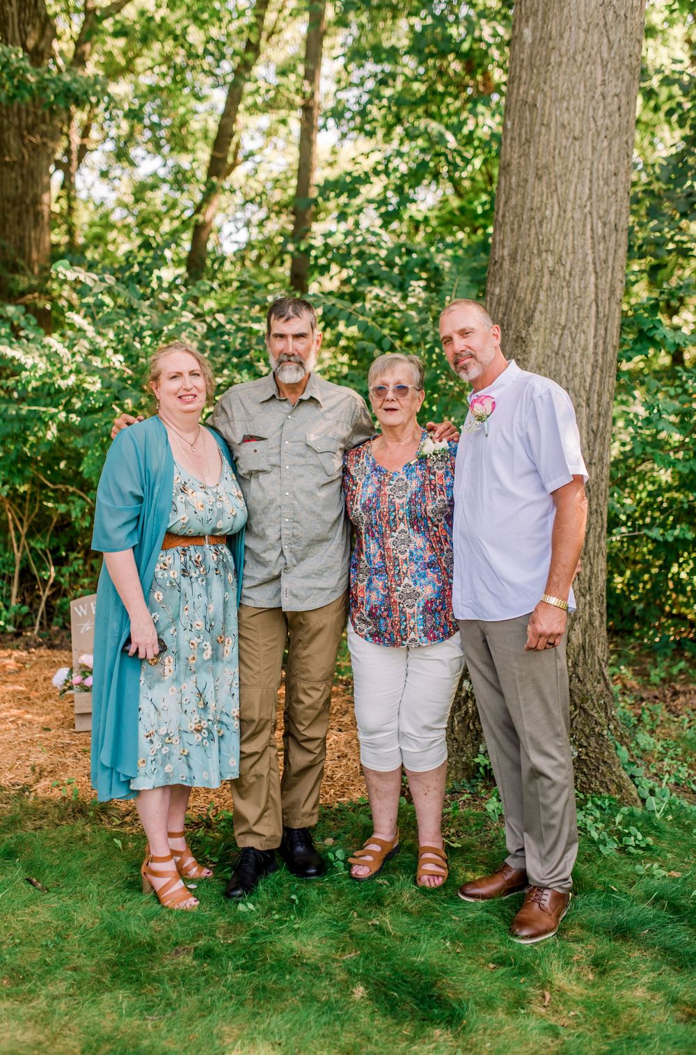 Four people posing outdoors near trees at a summer gathering, dressed in casual semi-formal attire.