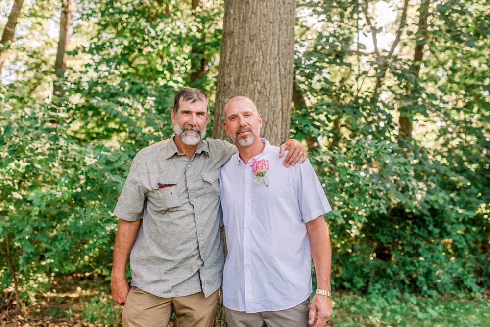 Two men smiling together outdoors near a large tree, one wearing a floral boutonniere, likely at a wedding celebration.