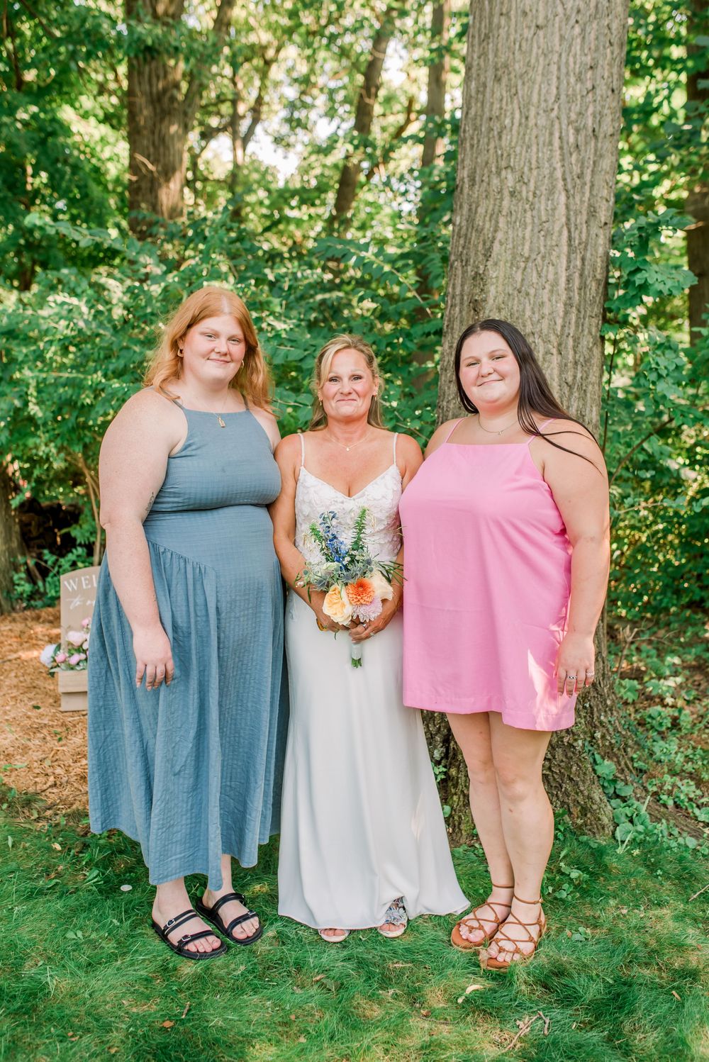 Bride in white gown holds bouquet flanked by two smiling women in blue maxi dress and pink mini dress outdoors.