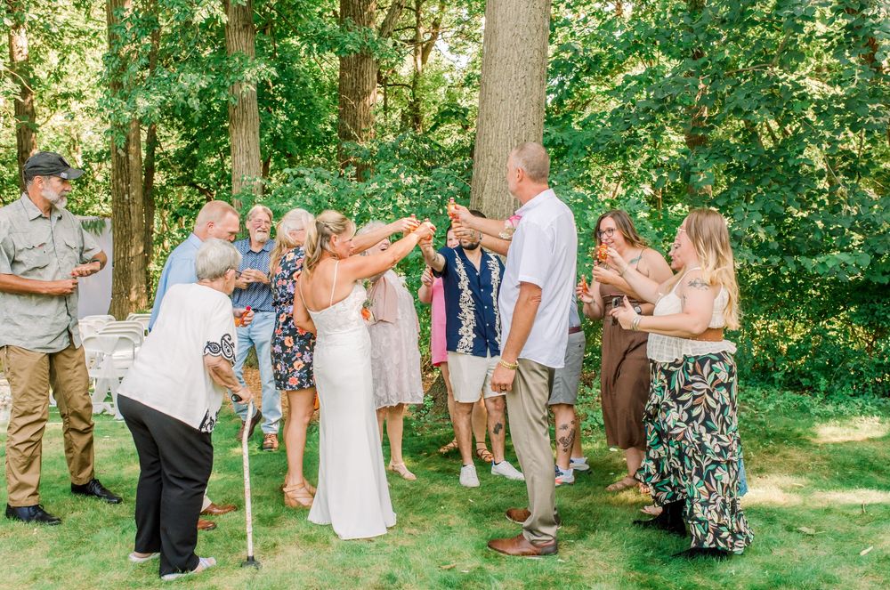 Bride and groom celebrate outdoors with guests raising glasses in a sunny backyard wedding toast surrounded by trees.