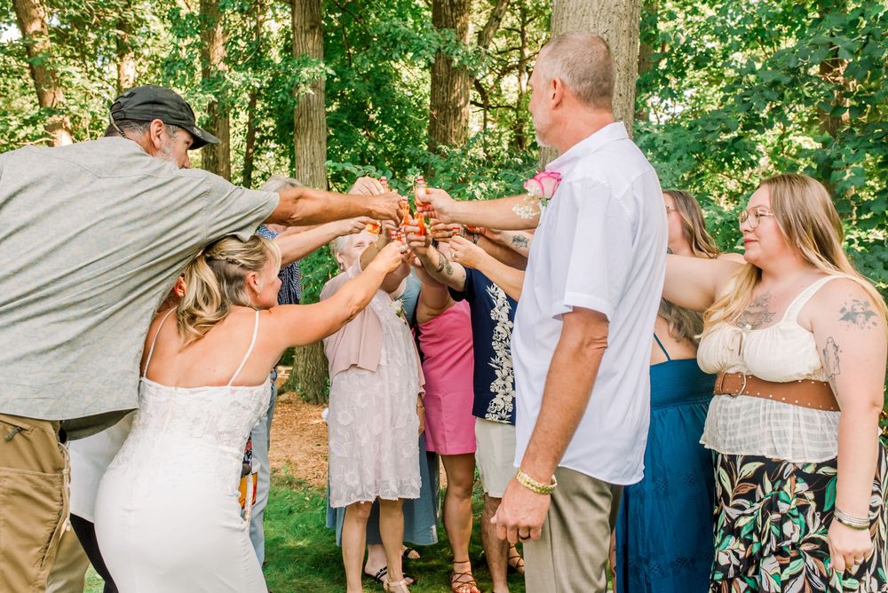 Wedding guests raising glasses in a celebratory toast outdoors among lush green trees at a summer wedding reception.