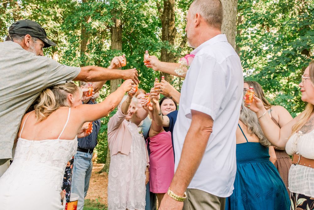 Wedding guests raising hands in celebration outdoors, bride in white dress surrounded by joyful attendees under green trees.