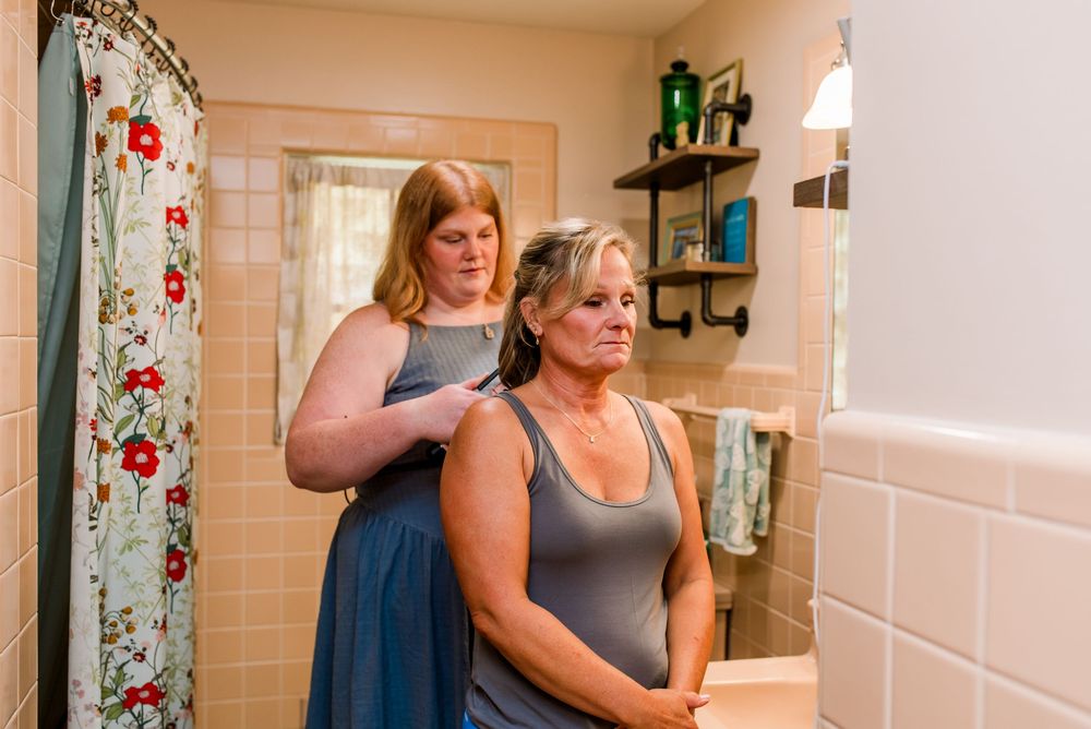 Woman styling another woman's hair in a bathroom with floral shower curtain and tiled walls.