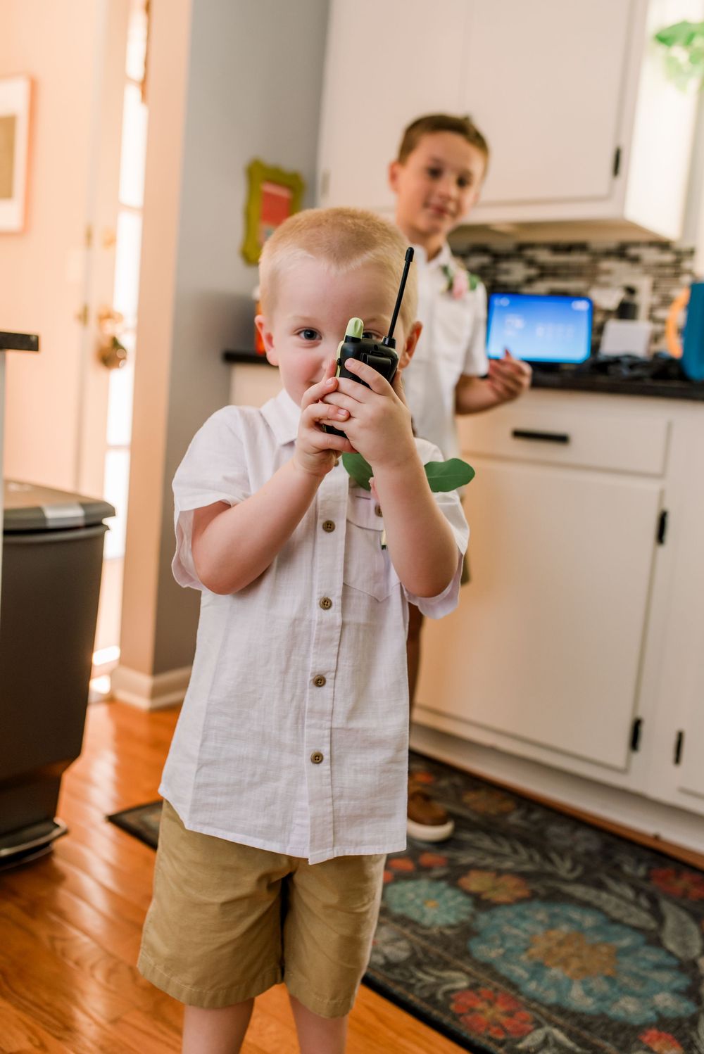Young boy in white shirt holds walkie-talkie indoors while older boy stands in kitchen background.