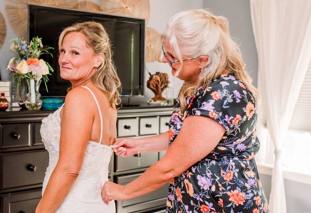 A bride in a white lace wedding dress gets help fastening her gown from an older woman in a floral dress.