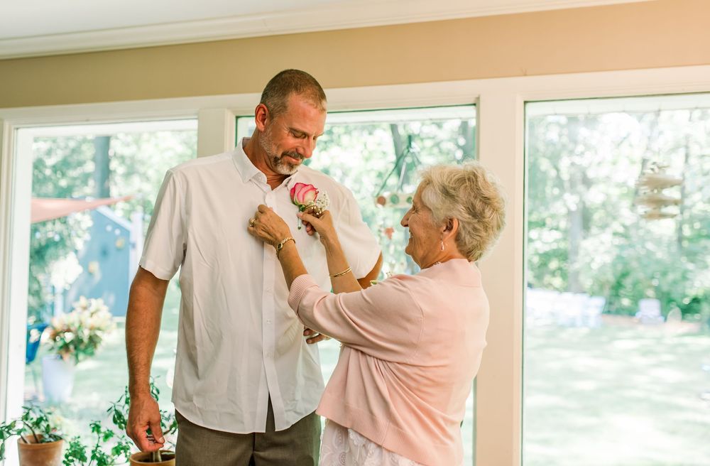 Elderly woman pinning a boutonniere on a tall man's white shirt indoors near large windows.