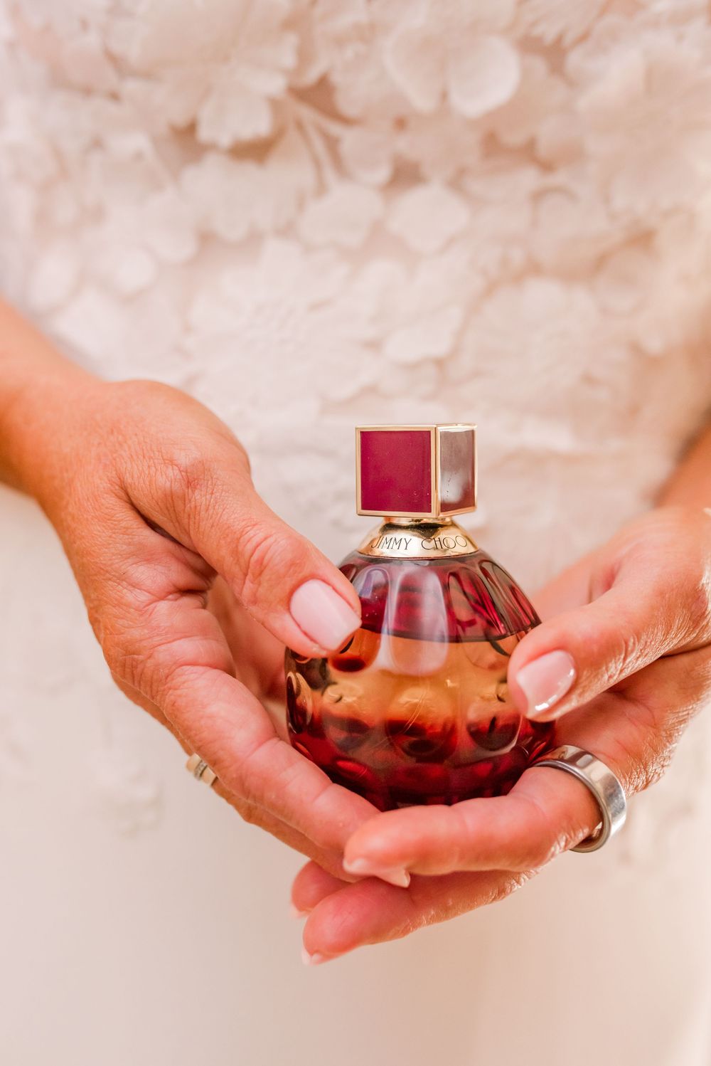 Bride holding a red and gold Jimmy Choo perfume bottle with manicured nails against a white floral dress.