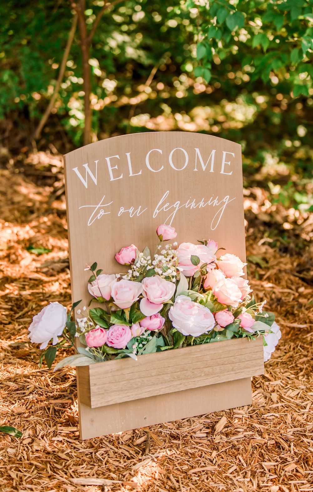 Wooden welcome sign with pink roses in a planter box, reading Welcome to our beginning, set outdoors.
