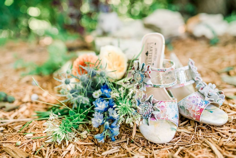 Bridal heels with floral bouquet featuring blue and peach flowers on rustic hay ground at outdoor wedding.