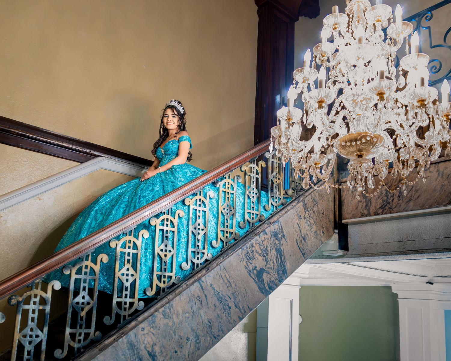 Young woman in teal quinceañera gown poses gracefully on ornate staircase beside elegant crystal chandelier.
