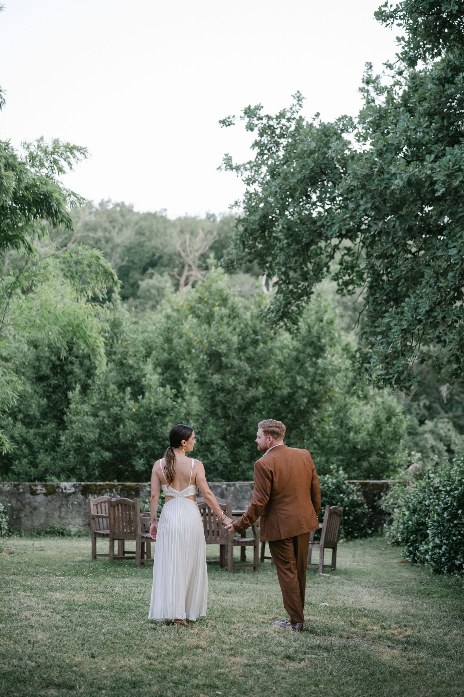 A bride and groom hold hands walking through a lush garden, she in a white gown, he in a brown suit.