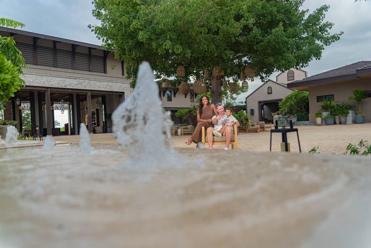 Family sitting on a bench near a fountain in a serene courtyard with lush trees and elegant buildings.