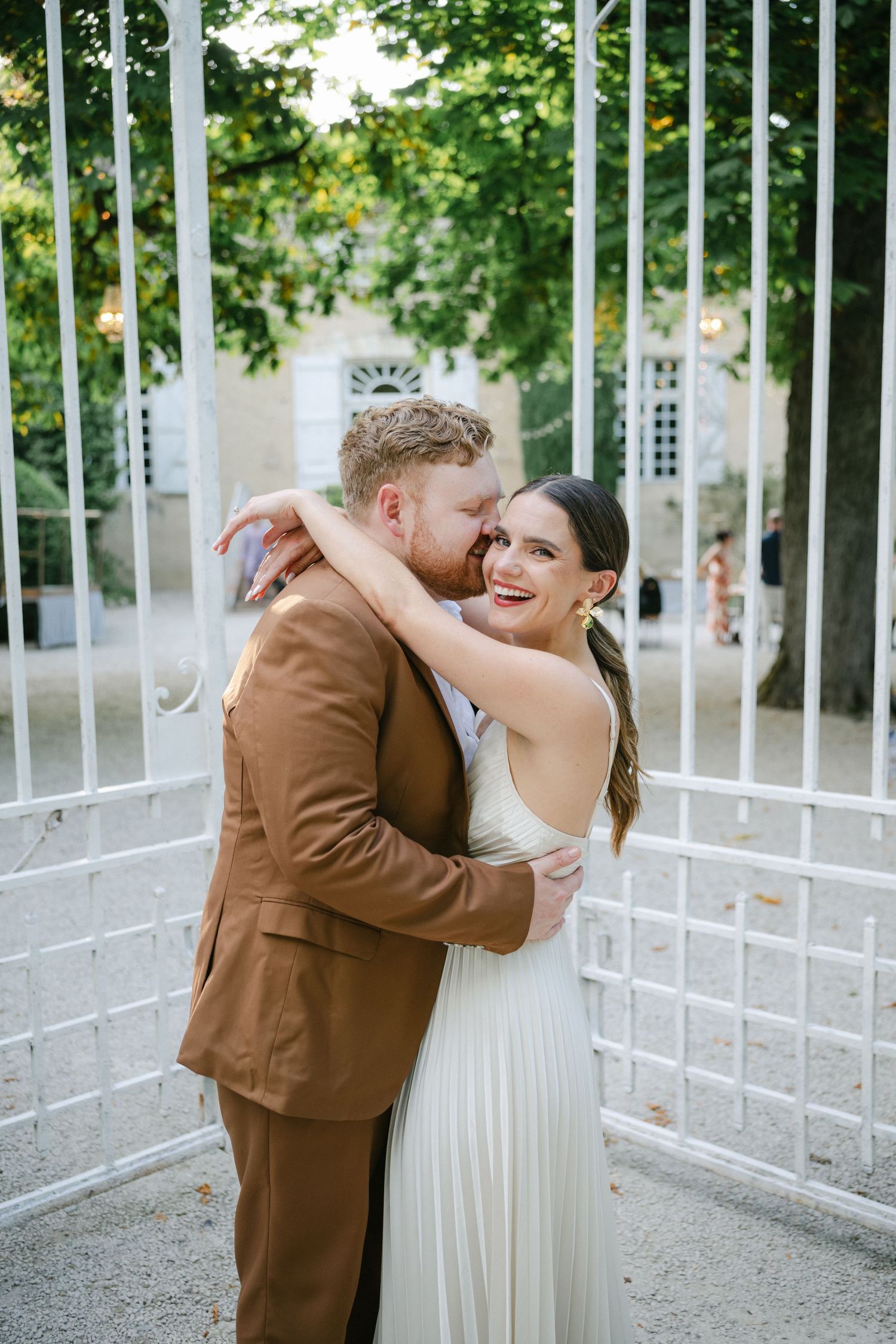 A smiling bride in a white dress sits on stone steps with a groom in a brown suit embracing her tenderly.
